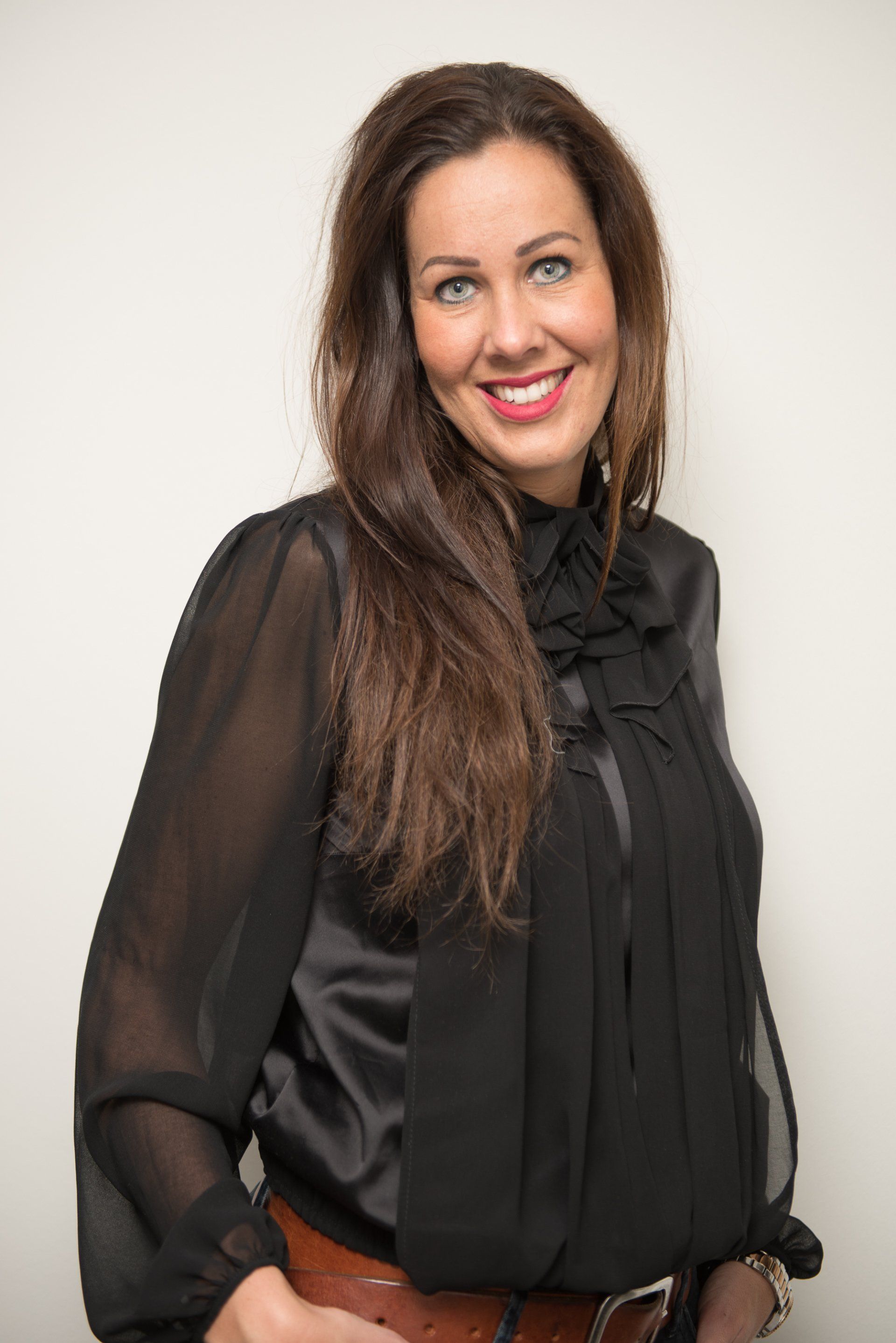Een persoon met lang bruin haar, gekleed in een zwarte blouse met lange mouwen, lacht naar de camera tegen een neutrale achtergrond.