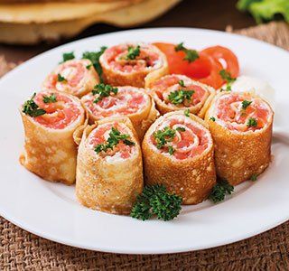 Een bord hartige crêpes gevuld met gerookte zalm en roomkaas, gegarneerd met verse peterselie en sch schijfjes tomaat.