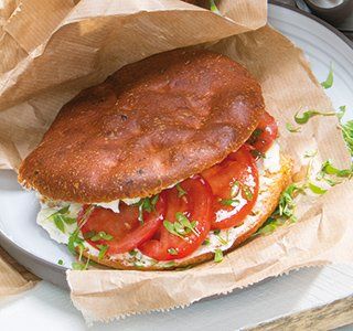 Een sandwich op een met bruin papier bekleed bord, met geroosterde broodjes, plakjes tomaat, roomkaas en wat microgroenten.