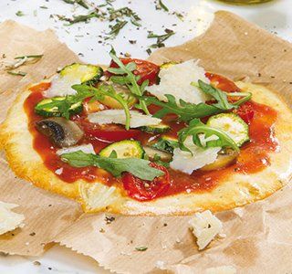 Een kleine pizza op bakpapier, belegd met tomatensaus, plakjes courgette, champignons, rucola en geraspte kaas.