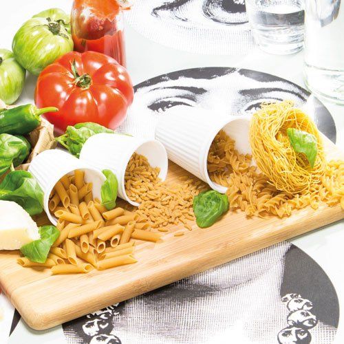 Houten plank met witte kopjes gevuld met verschillende soorten pasta, verse tomaten, paprika's en basilicum op een tafelblad met een patroon.