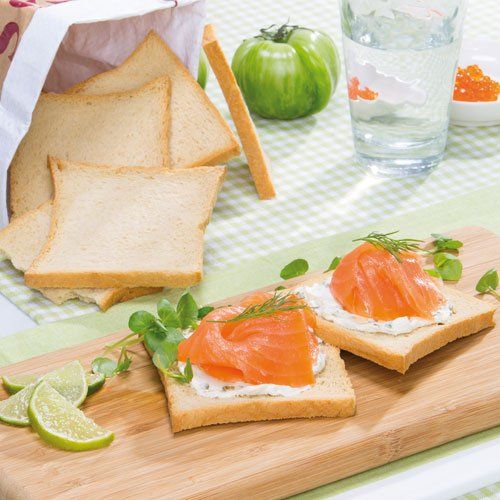 Twee sneetjes geroosterd brood belegd met roomkaas, gerookte zalm en kruiden, geserveerd op een houten plankje met schijfjes limoen en een groene tomaat.