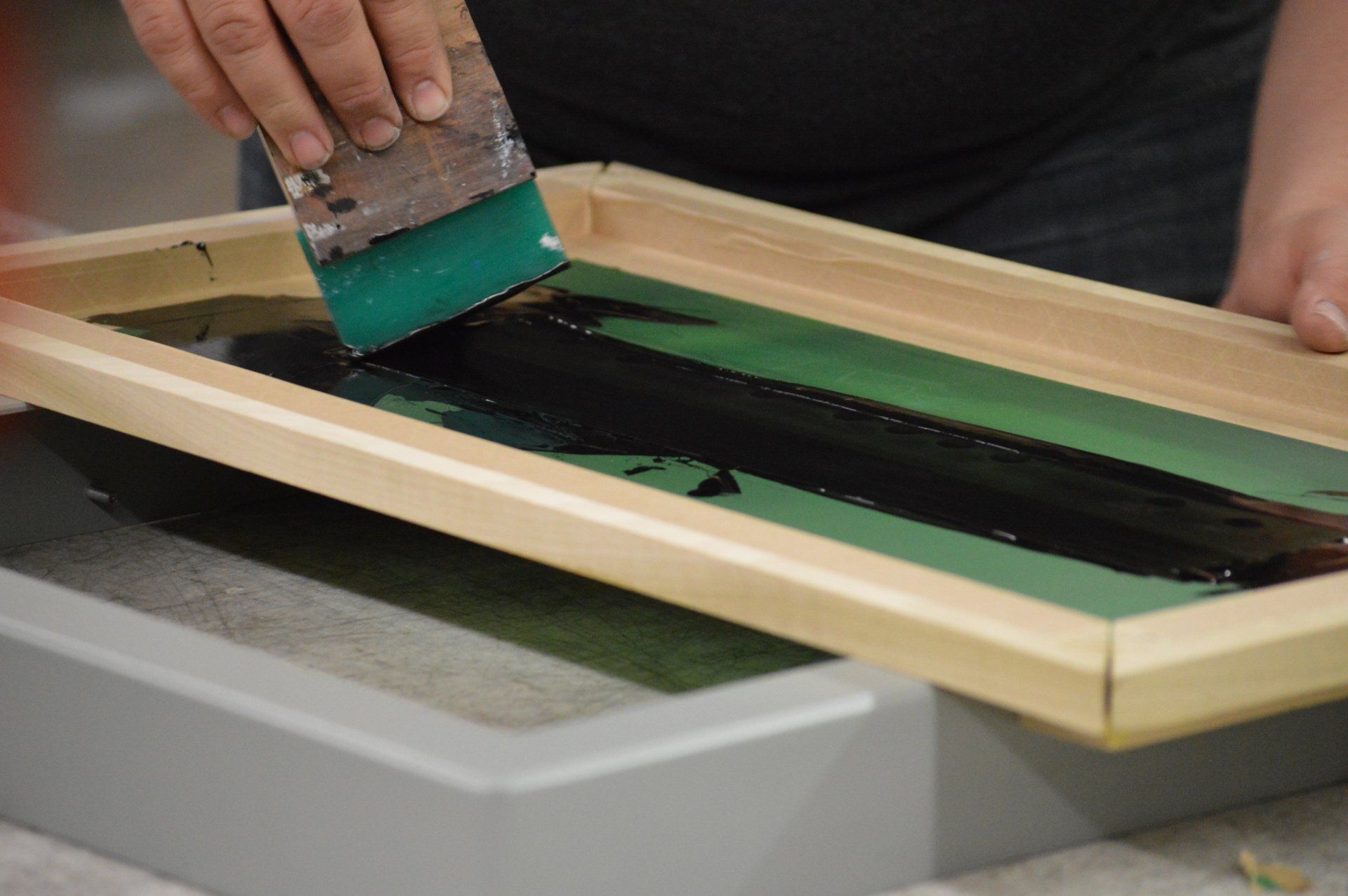 A person is using a green spatula to spread paint on a screen