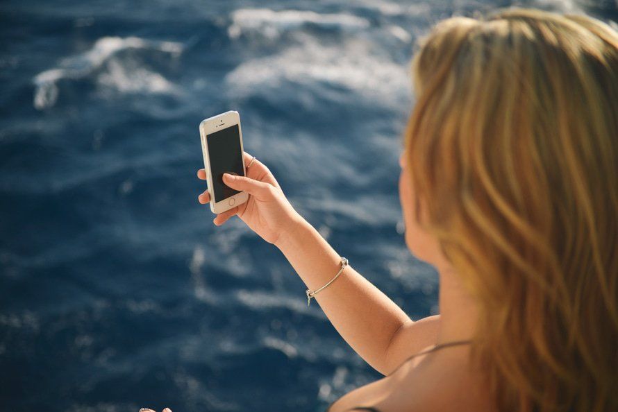 Person holding a smartphone above blue ocean water, taking a photo from a boat
