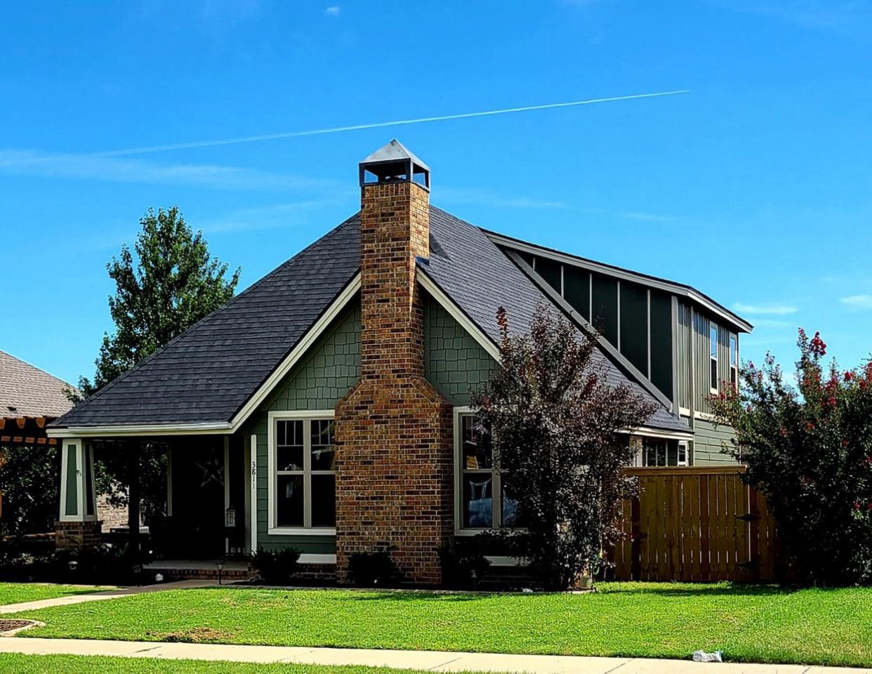 Suburban brick house with a steep dark roof, chimney, and green lawn under a blue sky
