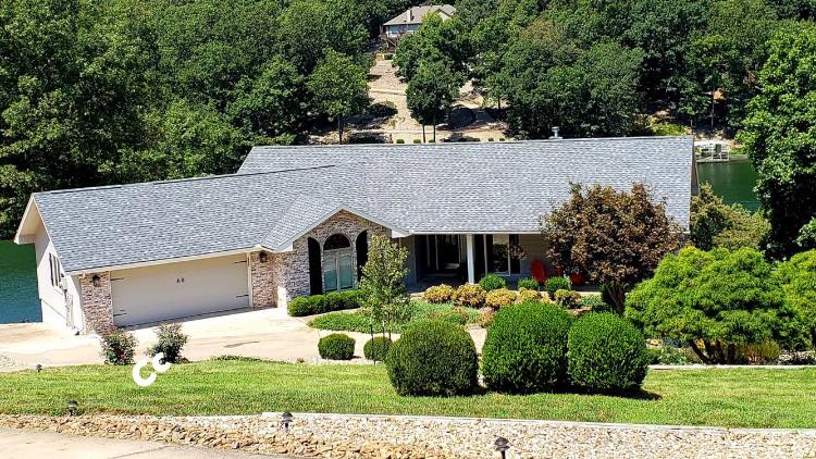 Single-story suburban house with gray roof, stone facade, and landscaped front yard on a wooded hillside