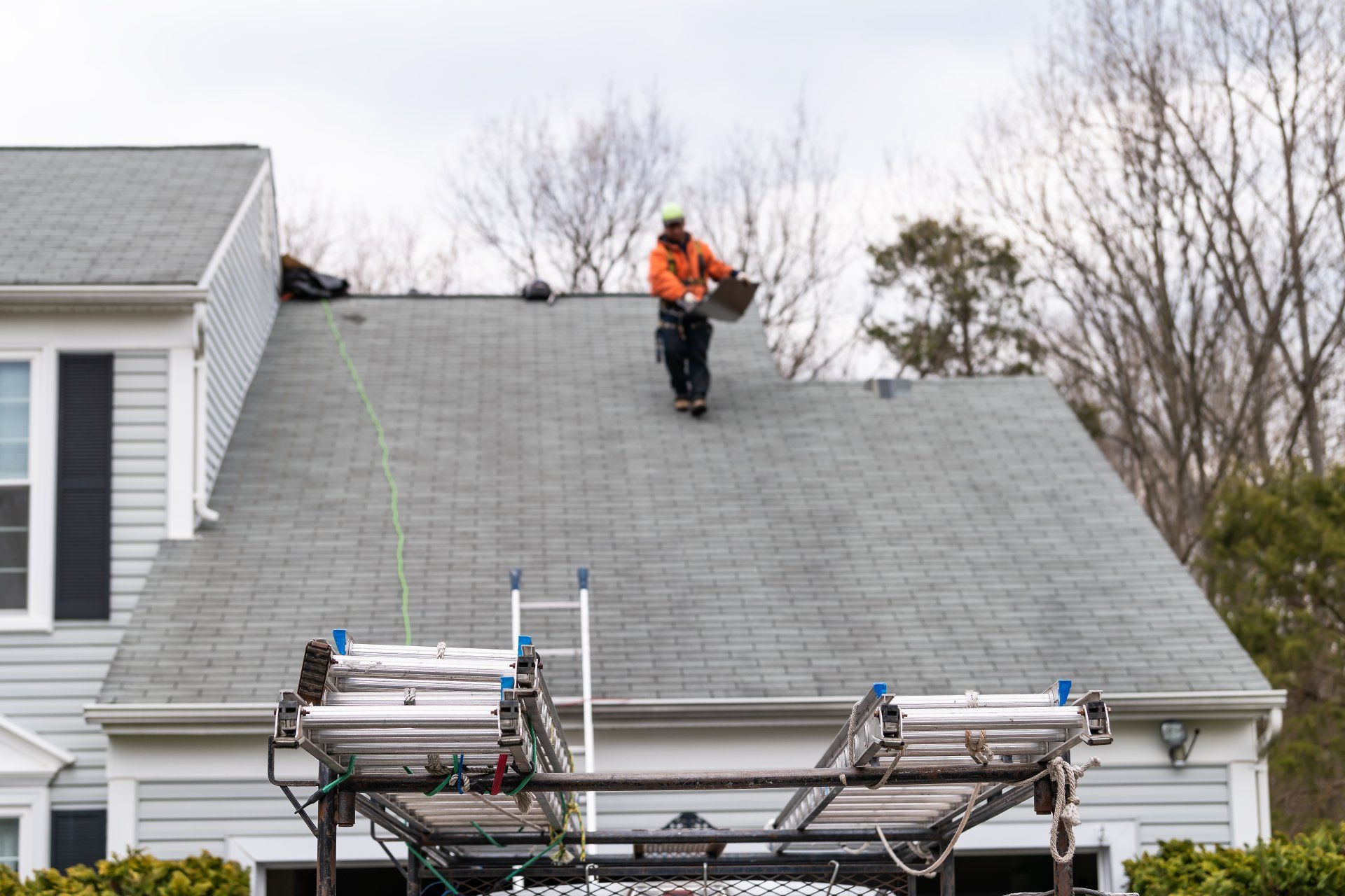 Worker in orange safety vest on a house roof, with a roof rack or equipment below.