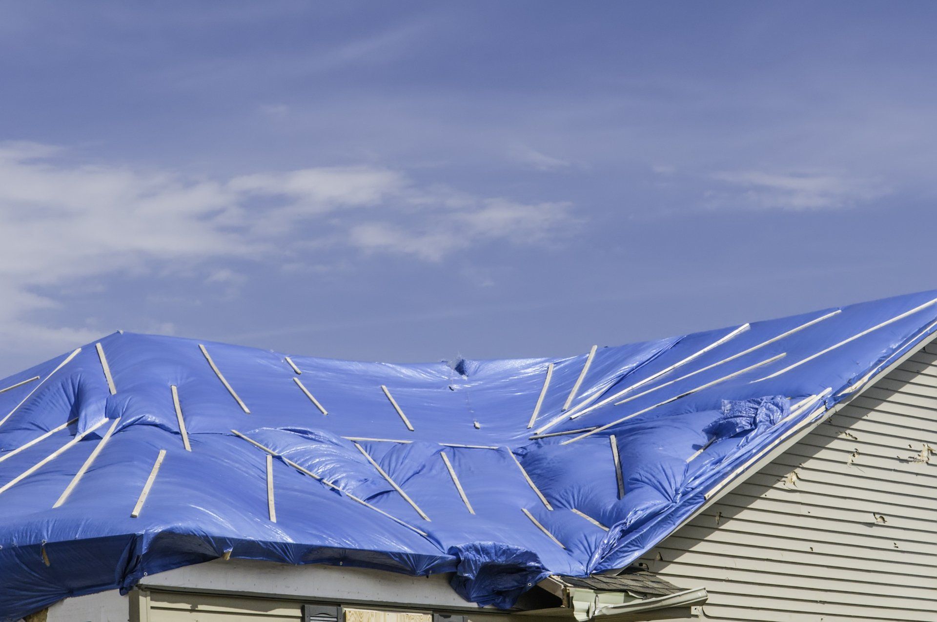 Blue tarp covering a damaged roof with torn shingles under a cloudy sky