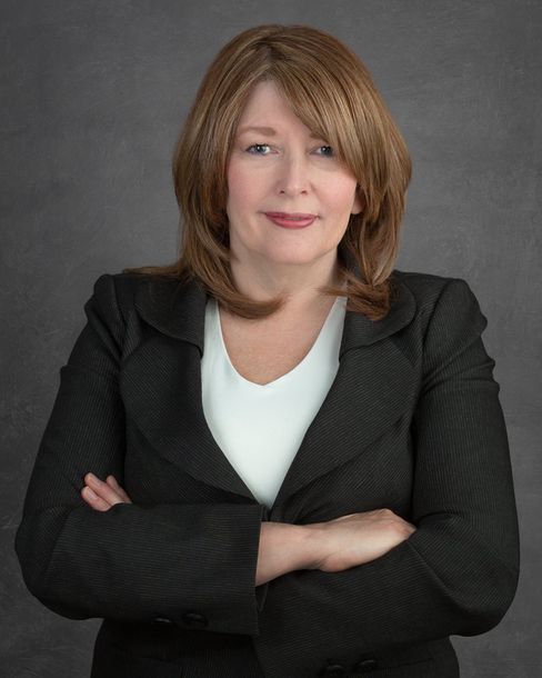 A woman in a black jacket and white shirt is standing with her arms crossed.