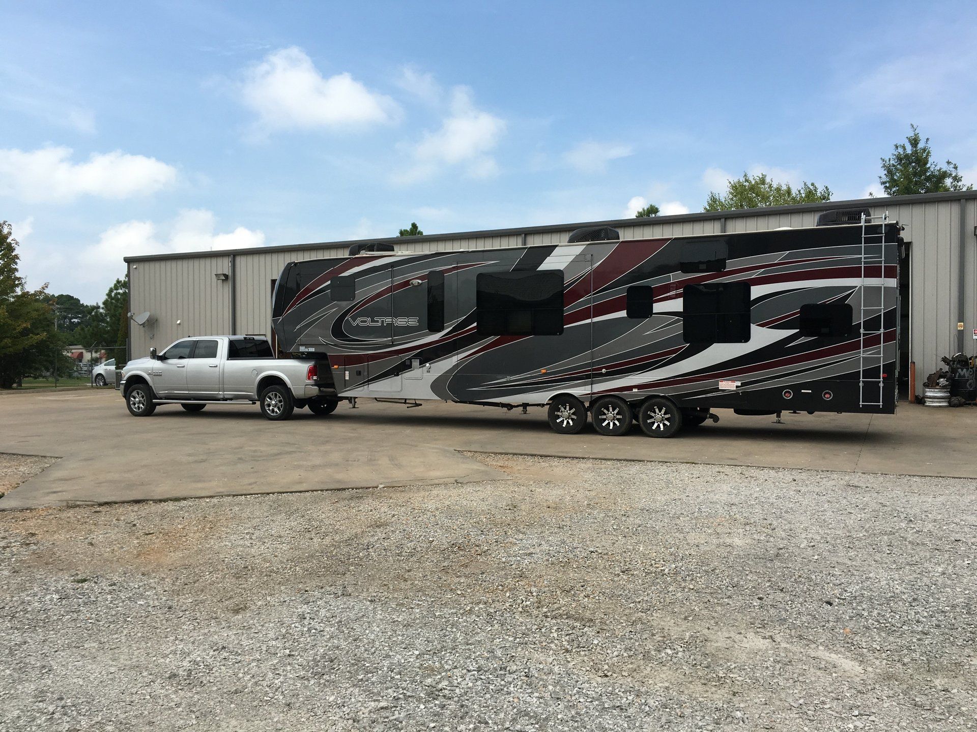 Silver truck towing a large black, gray, and red toy hauler trailer in front of a building on a sunny day.