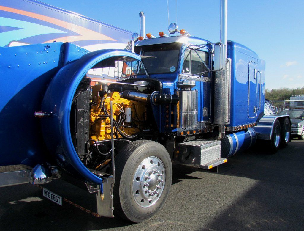 Blue semi-truck with open hood revealing the yellow engine. Parked outdoors on a sunny day.