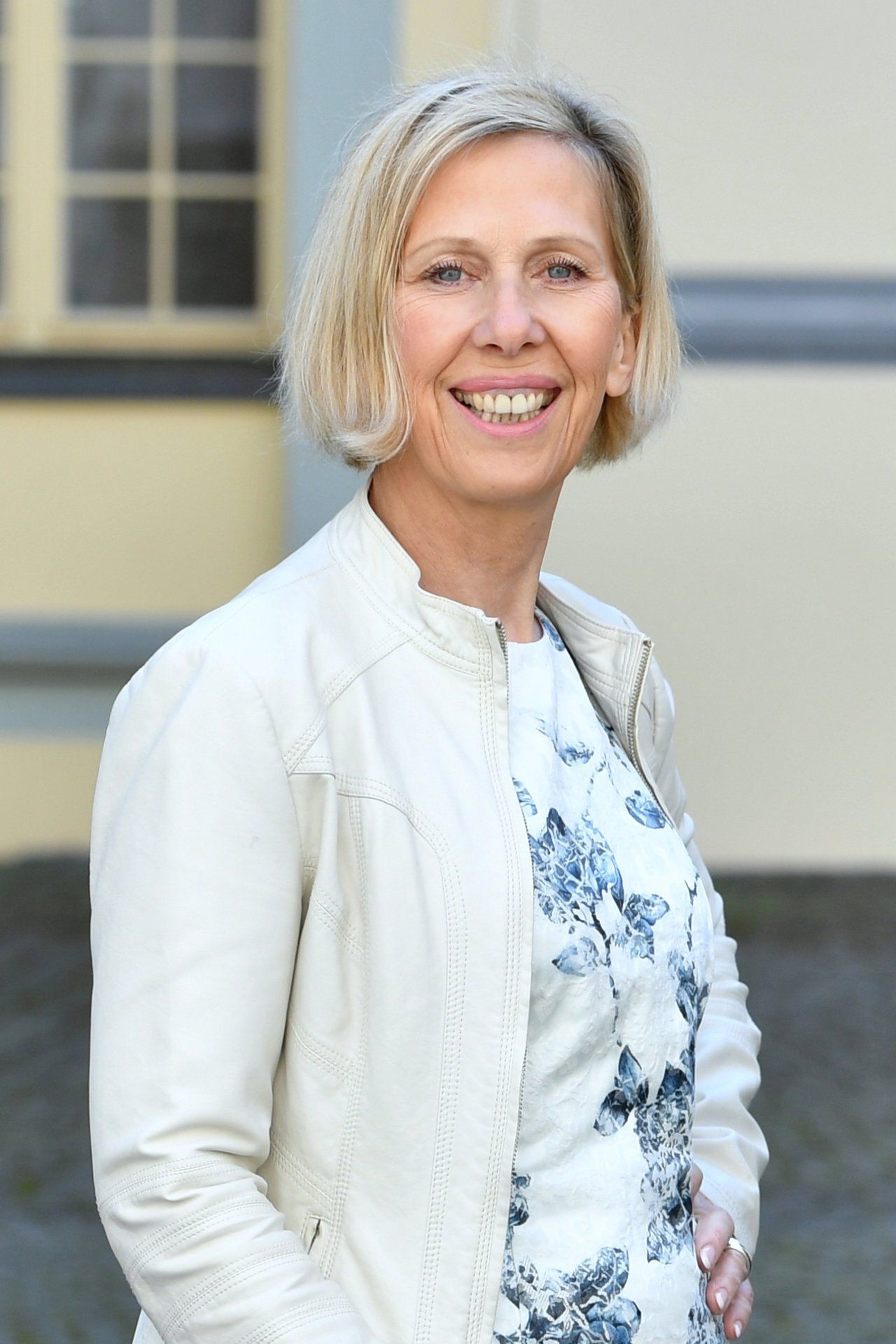 Eine Frau in einer weißen Jacke und einem geblümten Kleid lächelt in die Kamera.