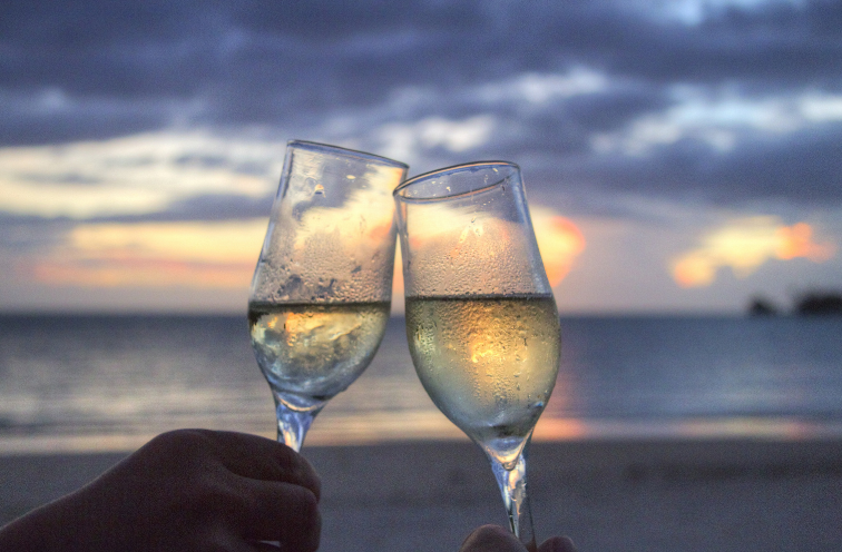 Two champagne flutes clink on a beach at sunset with the ocean in the background
