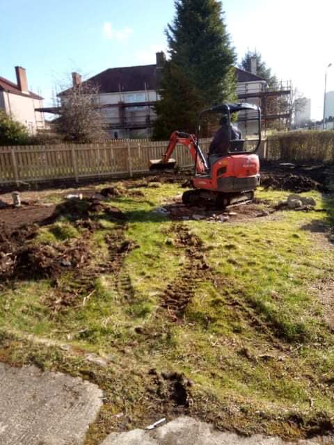 A small red excavator digging a muddy backyard beside a fence and house.