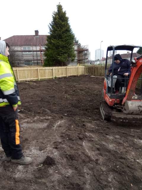 Construction workers operating a mini excavator on a muddy lot beside a fenced house and evergreen tree