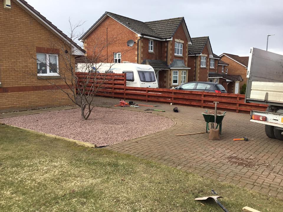 Brick house backyard with paved patio, caravan, lawn, and wooden fence under a gray sky