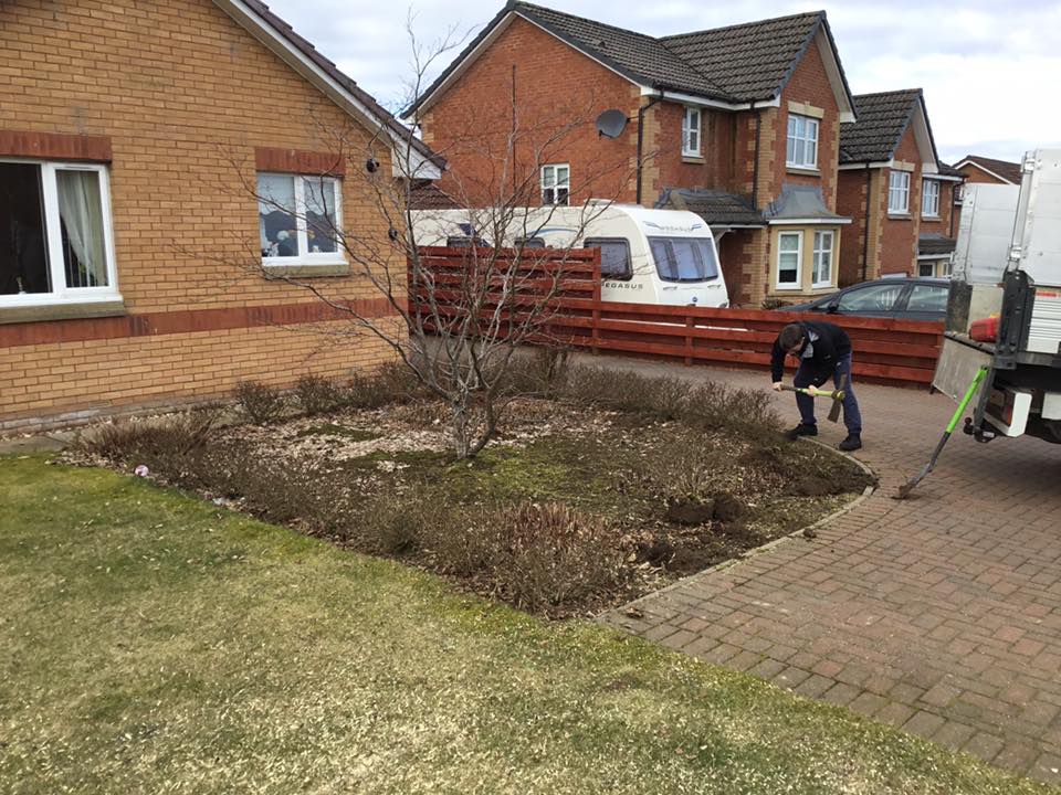 Person bending over a front-yard garden bed beside a brick house and parked van