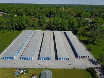 Aerial view of a storage facility with multiple rows of units and a concrete driveway surrounded by trees.