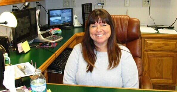 Woman smiling at a desk in an office setting. She has dark hair and wears a gray sweater.