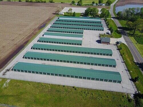 Overhead view of a storage facility with multiple rows of green-roofed units on a concrete lot, surrounded by fields and a road.