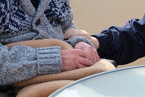 Person in gray sweater and seated, hands clasped by another person's hand in a blue sleeve, both over a brown blanket.