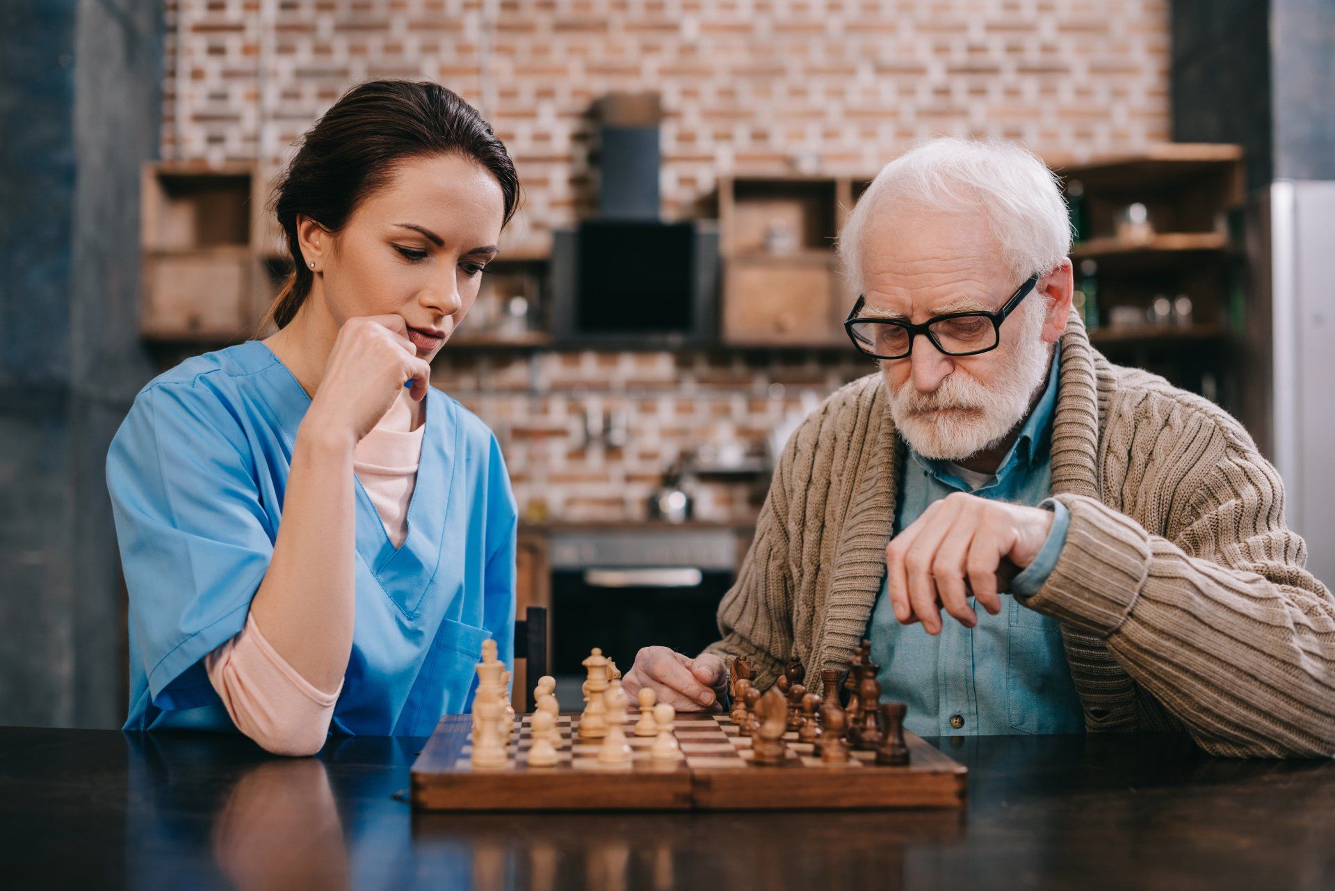 Woman in blue scrubs watches intently as an older man with glasses moves a chess piece at a table.