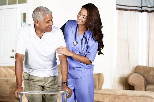 A young healthcare worker in blue scrubs helps an elderly man walk with a walker in a home setting; both are smiling.