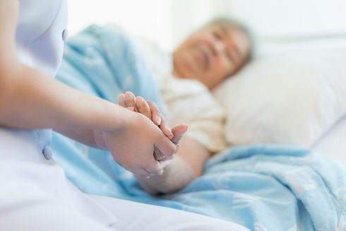 Nurse holding the hand of an elderly patient lying in bed, offering support and comfort. Soft blues and whites, indoor setting.