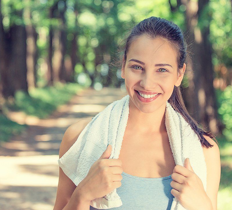 Woman smiling, holding a towel around her neck, standing on a path in a park with trees in the background.