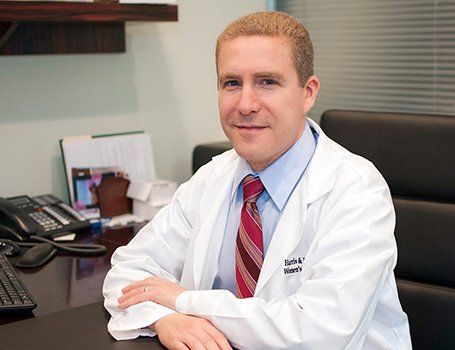 Man in a lab coat, seated at a desk, looking at the camera. He wears a tie and smiles. Office setting.
