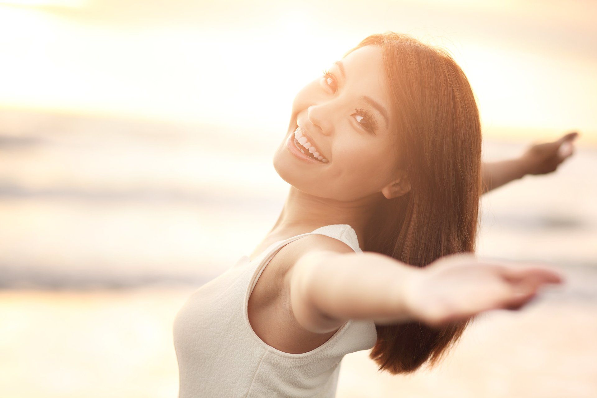 Woman with arms outstretched, smiling at the camera on a beach at sunset.