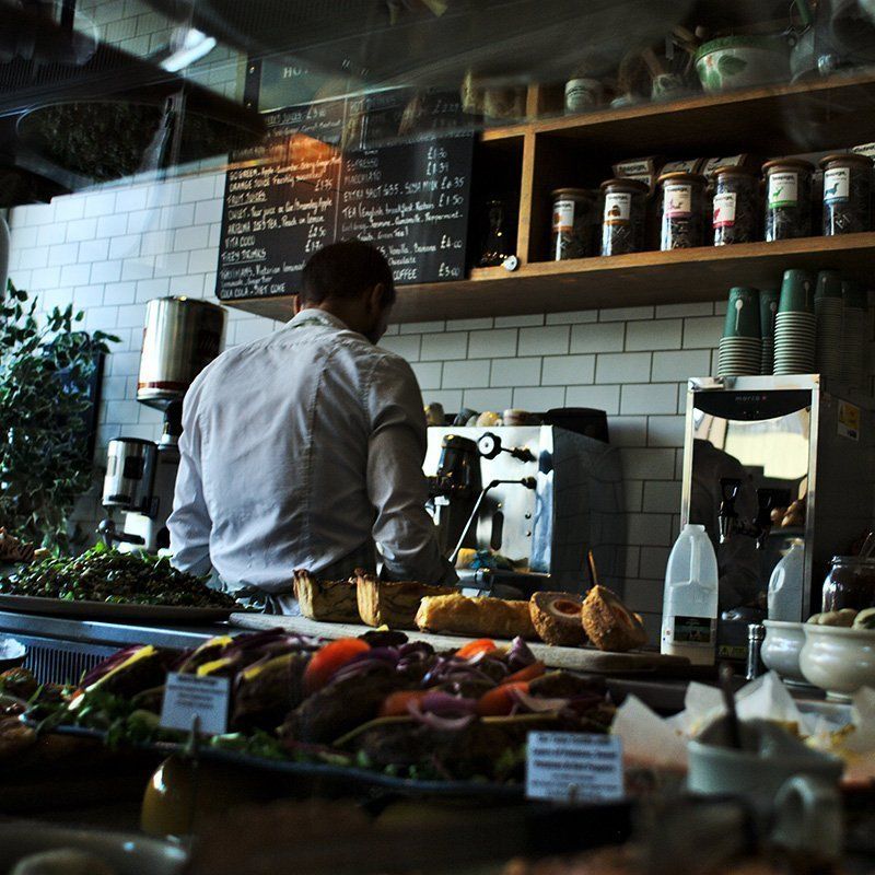 Man behind a cafe counter preparing a coffee, food display in front, menu and shelves of goods behind.