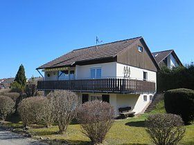 Ein weißes Haus mit einem braunen Dach und einem Balkon, umgeben von Büschen und Bäumen.