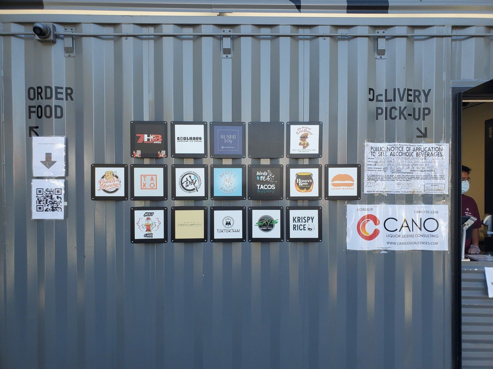 Shipping container with square framed signs, order food and delivery pick-up signs, and a person behind a counter.