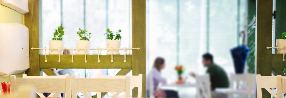Interior of a cafe with a window. People seated at a table, with plants on a shelf in front.