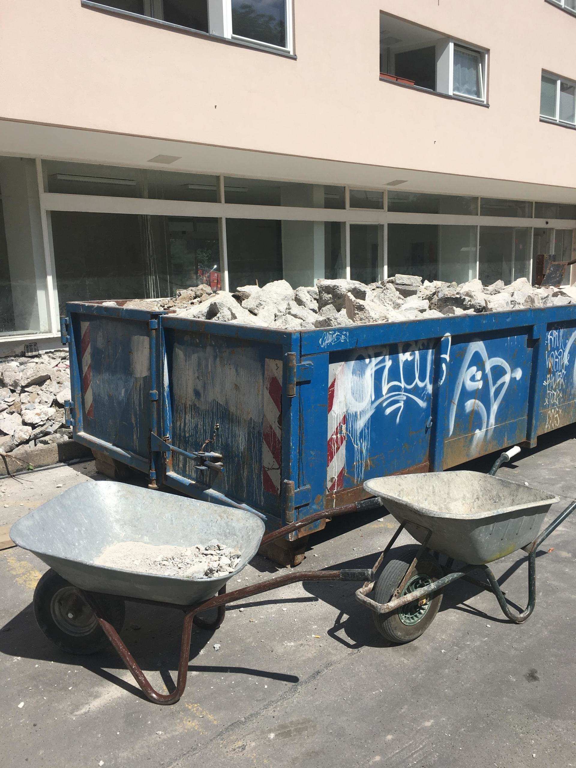 Blauer Müllcontainer, der mit Schutt überfüllt ist, zwei Schubkarren davor, in der Nähe eines Gebäudes.