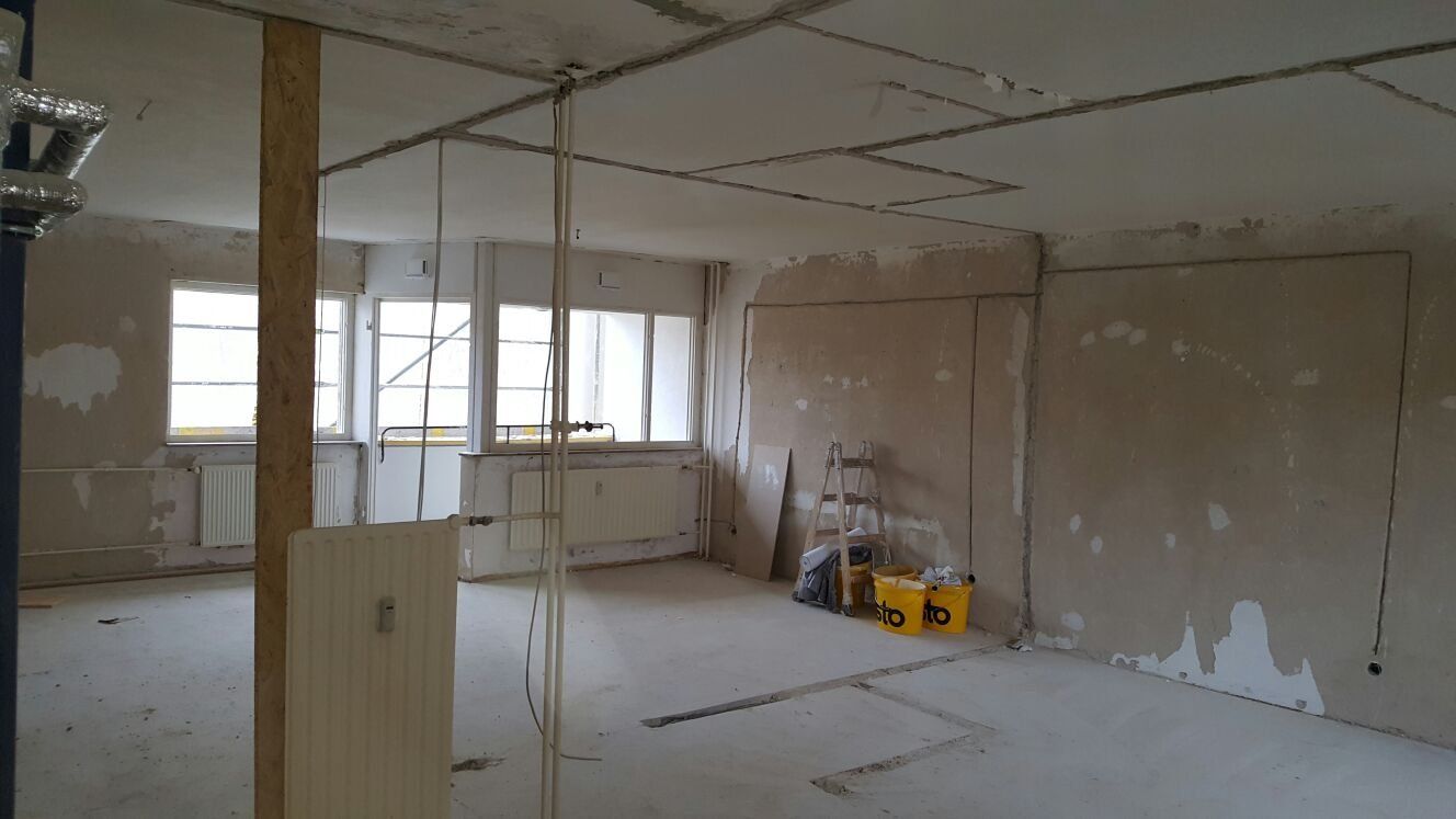 Im Bau befindlicher Raum mit kahlen Wänden, freiliegenden Kabeln, Fenstern und Baumaterialien.