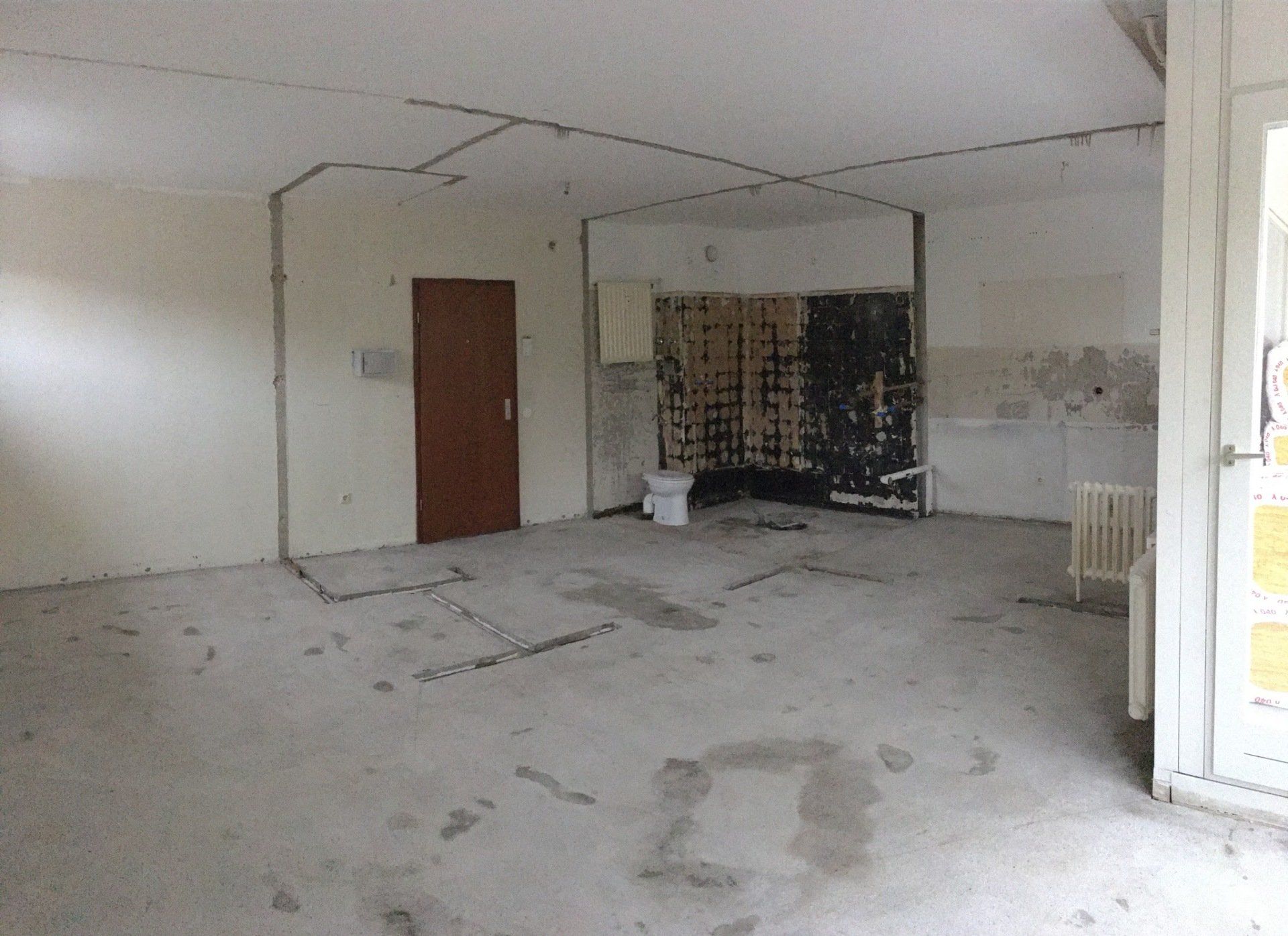 Ein leerer, staubiger Raum wird gerade renoviert; Wände, Decke und Boden sind grau und abgenutzt. Links ist eine braune Tür.