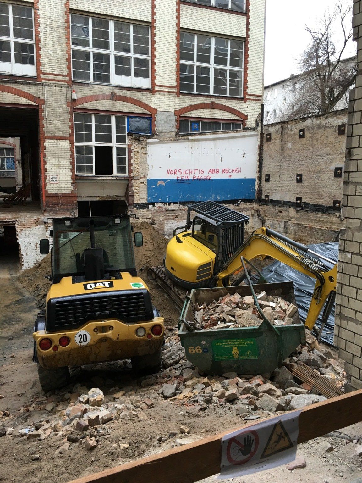 Baustelle: Ein gelber Bagger lädt Schutt in einen grünen Müllcontainer, in der Nähe steht ein gelber Gabelstapler.