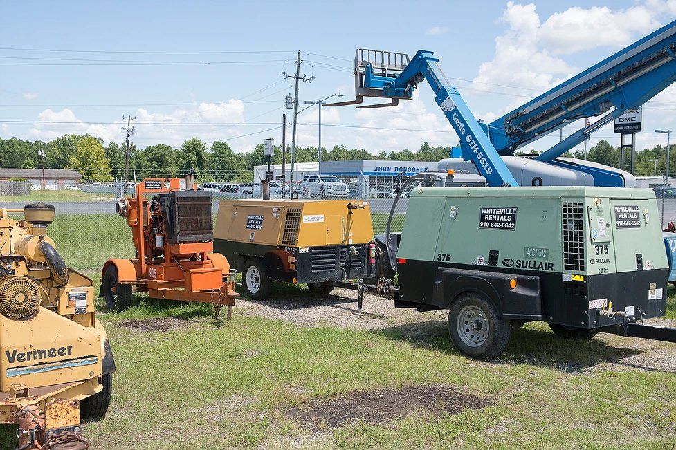 Assortment of construction equipment on a grassy lot; orange asphalt machine, generators, and a blue lift.