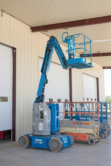 Blue Genie articulating boom lift extended, parked next to other lifts inside a building.