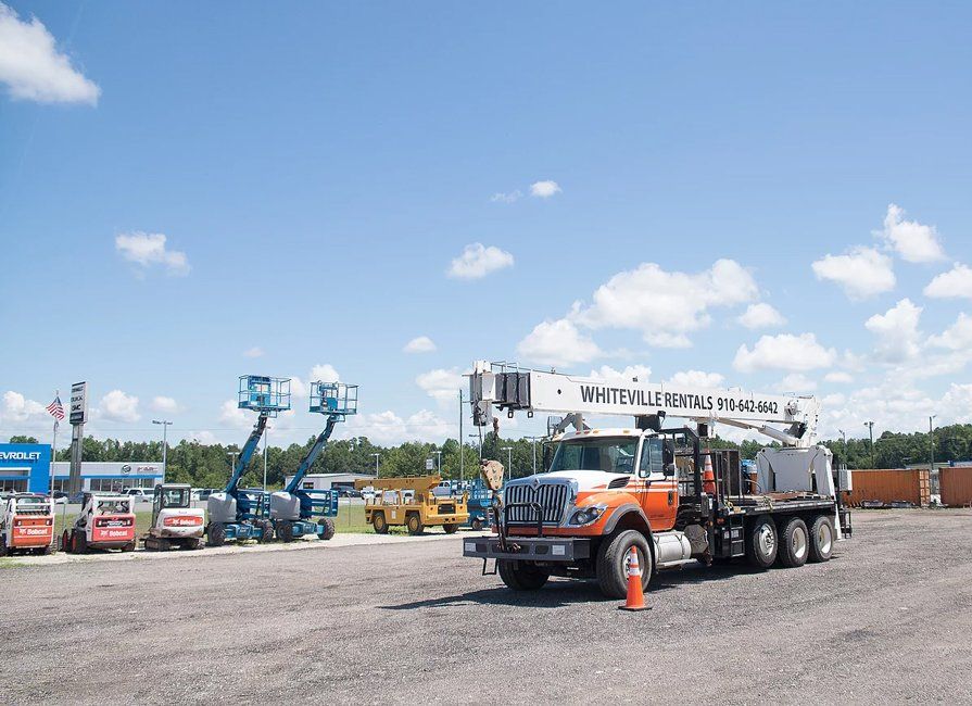 Truck with a crane, parked in a lot with construction equipment under a blue sky with clouds.