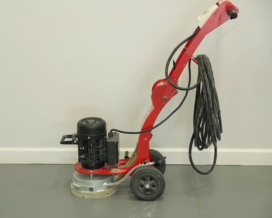 Red floor grinding machine on a grey floor, next to a pale wall, with a black power cord.