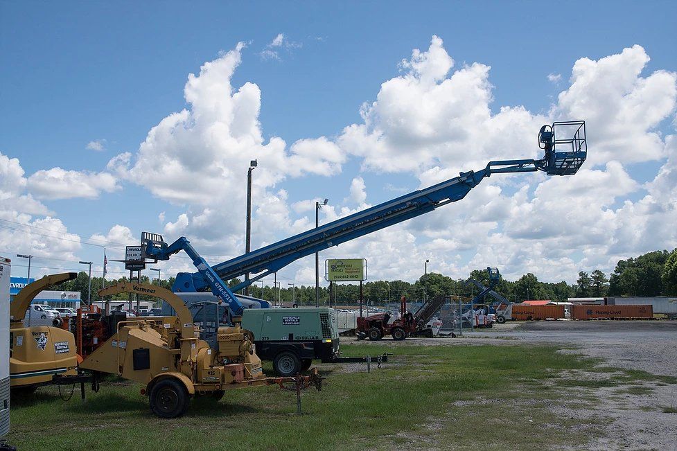 A blue aerial lift extends towards a bright, cloudy sky in a lot with equipment.