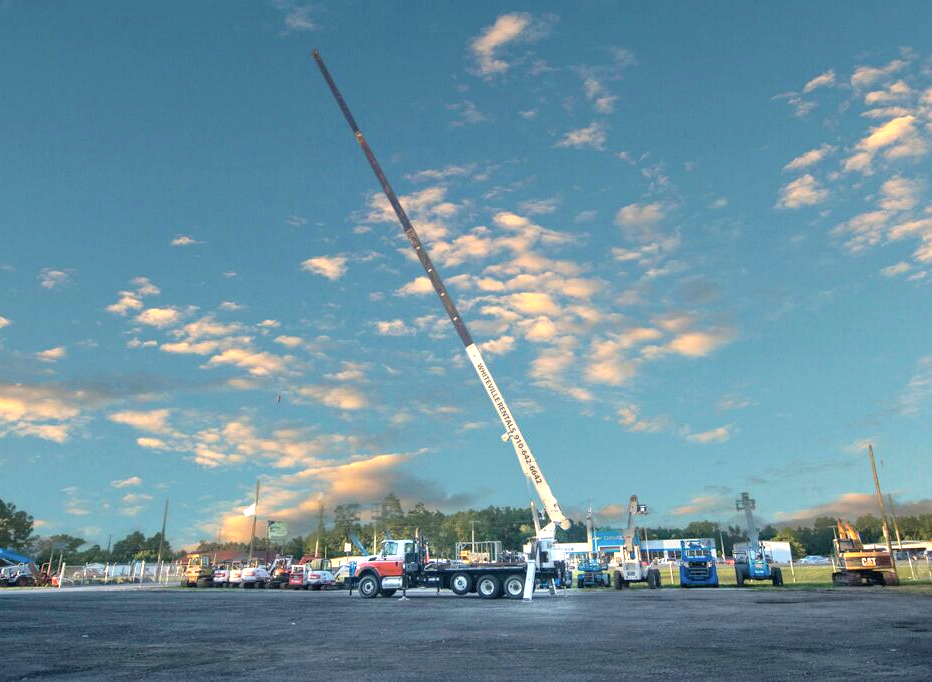A tall crane on a flatbed truck is raised against a blue sky with clouds, parked in a lot.