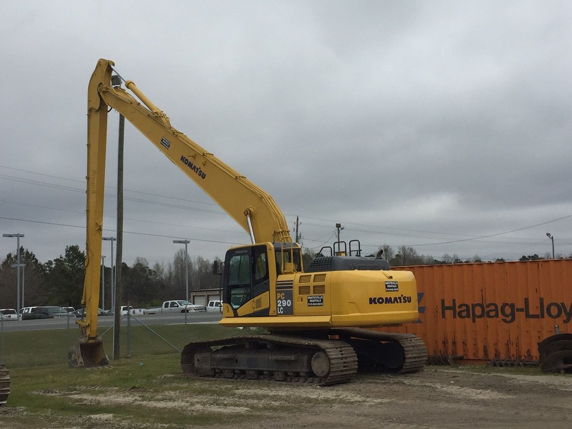 Yellow Komatsu excavator with long arm digging near a shipping container under an overcast sky.