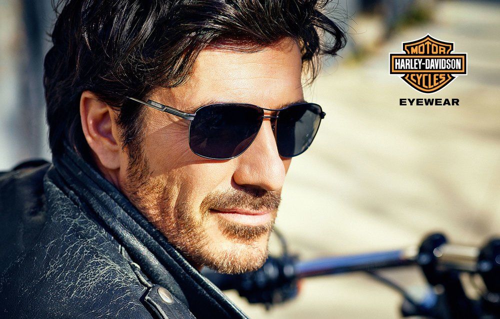 Man wearing sunglasses and a black leather jacket near a motorcycle, looking off to the side.