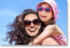 Woman and child smiling in sunglasses by the beach, with blue sky in the background