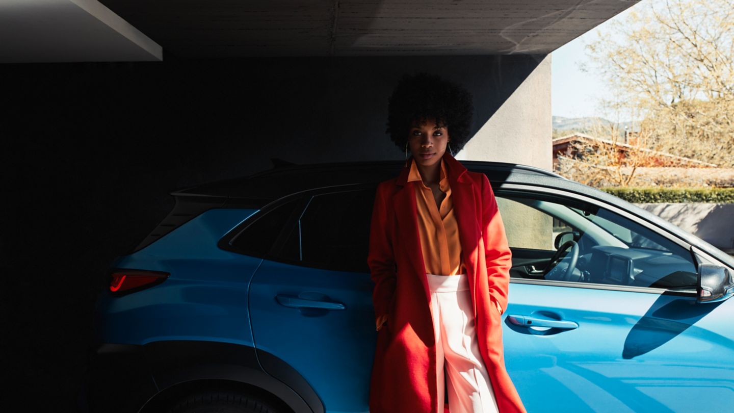 A woman in a red coat is standing next to a blue car.