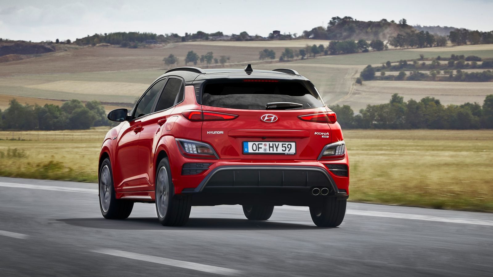 A red hyundai kona is driving down a road next to a field.